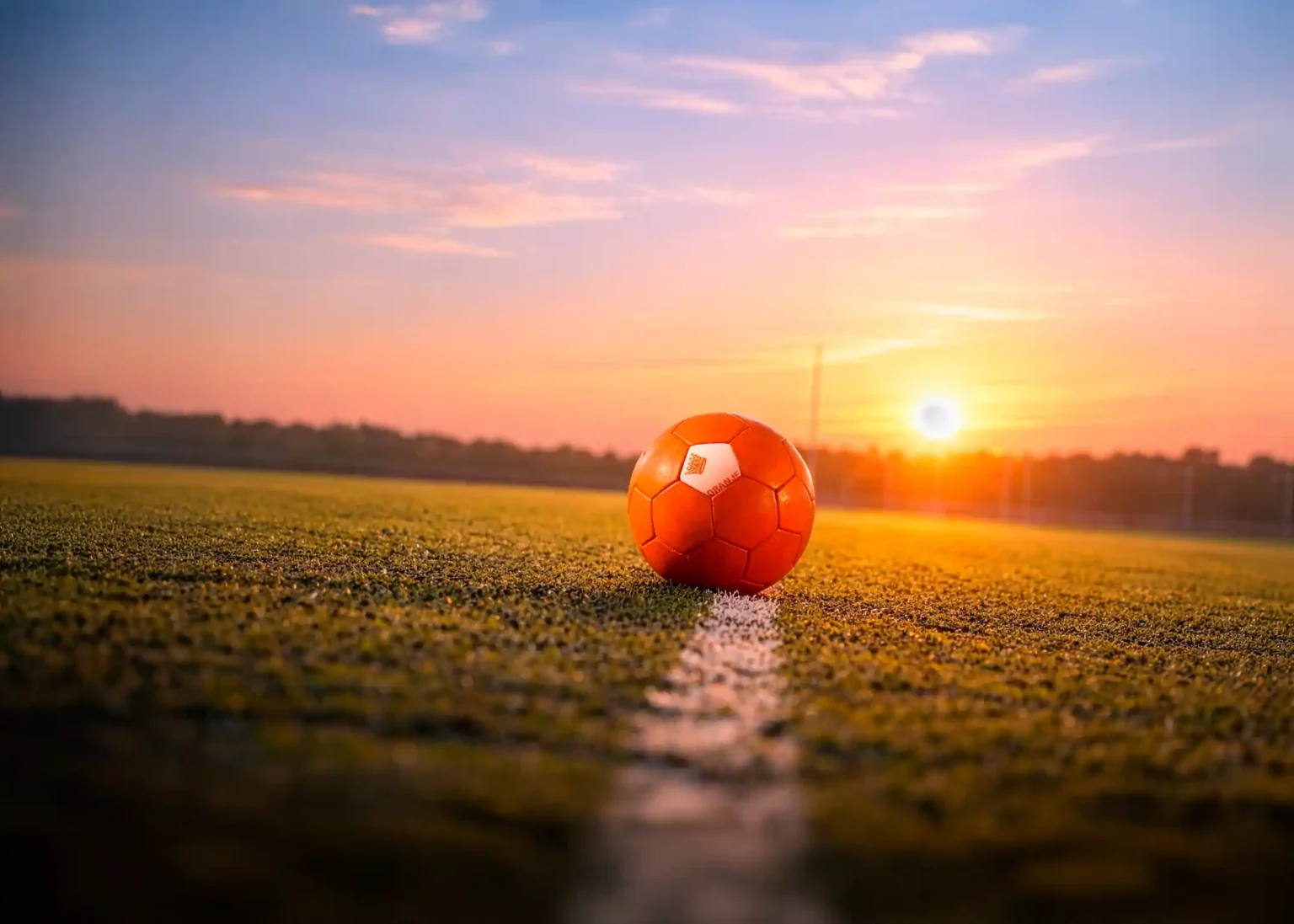 Orangefarbener Fußball auf der weißen Linie eines Rasenplatzes bei Sonnenuntergang, mit einem warm leuchtenden Himmel im Hintergrund